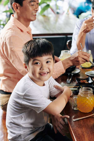 Portrait of cute smiling Asian boy sitting at dinner table with his family and looking at cameraの写真素材