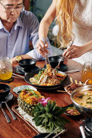 Housewife using fork and pliers when putting delicious Vietnamese dish from bowl in her plate and plate of her fatherの写真素材