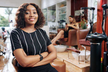 Portrait of beautiful confident young Black woman sitting in chair in her beauty salonの写真素材