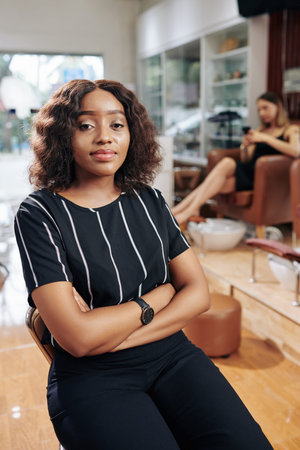 Pretty young Black woman sitting with arms folded and waiting for beautician in beauty salonの写真素材
