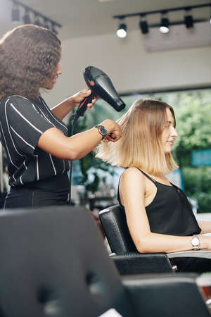 Stylist using professional dryer when blowdrying hair of young pretty woman in her salonの写真素材
