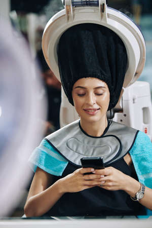 Smiling young woman texting friends and checking social media when receiving hair treatment in beauty salonの写真素材