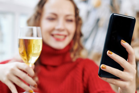 Smartphone in hand of smliing woman drinking wine on Christmas eve, selective focus