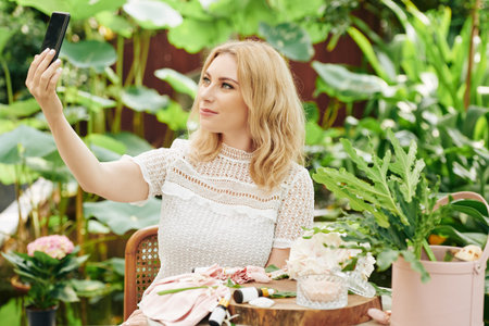 Attractive smiling young blond woman sitting outdoors with natural cosmetics on table in front of...