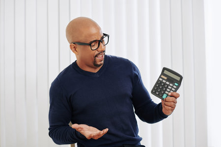 Frowning bald financial manager in glasses looking at calculator in his hand
