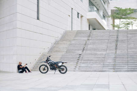 Black young man sitting on ground and taking photo of his motorcycle on smartphoneの写真素材