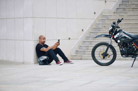 Muscular man in black clothes sitting on ground and texting friends after riding motorcycleの写真素材