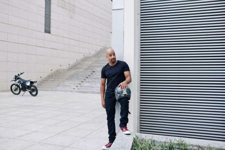 Handsome muscular man with helmet standing outdoors, his motorcycle in backgroundの写真素材