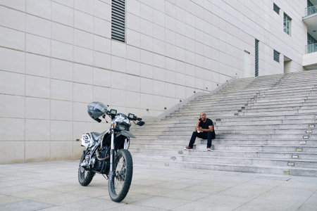 Smiling man sitting on steps, drinking coffee and talking on phone after riding on motorcycle