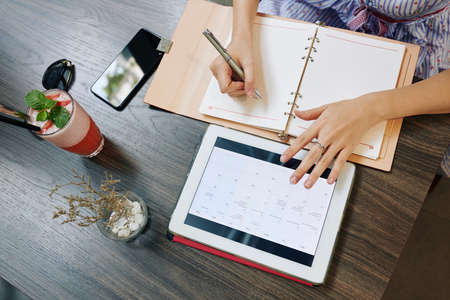 Hands of business lady checking calendar on tablet computer and writing in planner, view from aboveの写真素材
