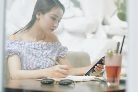 Serious pretty young Vietnamese woman checking text messages in her smartphone and writing in...