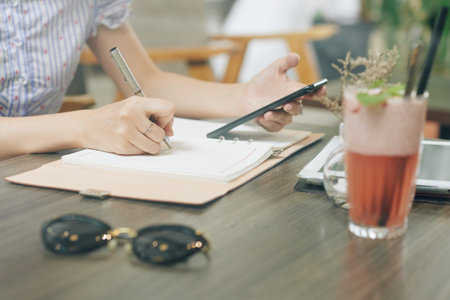 Hands of female businesswoman checking e-mails on her smartphone and writing down important thoughtsの写真素材