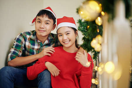 Portrait of happy hugging Asian teenage brother and sister in Santa hats sitting on steps and looking at cameraの写真素材