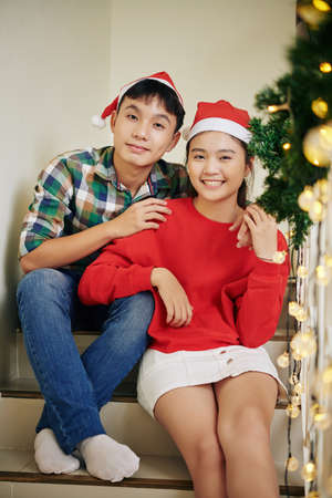 Smiling teenage boy hugging his sister in red sweater in Santa hat when they are resting on stairs on Christmas eveの写真素材