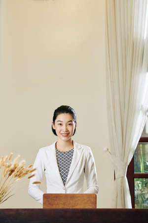 Portrait of young pretty Asian woman standing at reception with opened laptop and smiling at cameraの写真素材