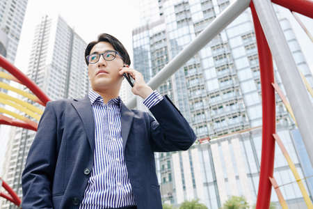 Serious investor standing in new city district with high buildings and talking on phoneの写真素材