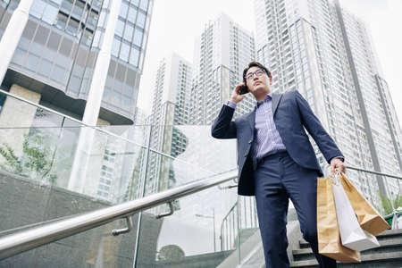 Serious Asian man in glasses carrying paper-bags when walking down the stairs and talking on phoneの写真素材