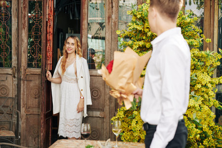 Smiling young woman opening door and looking at boyfriend waiting for her with bouquet of flowersの写真素材