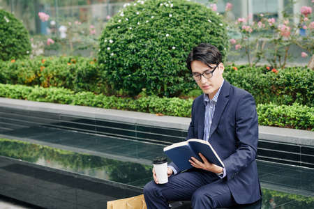 Intelligent Asian entrepreneur sitting on bench with cup of take away coffee and reading bookの写真素材