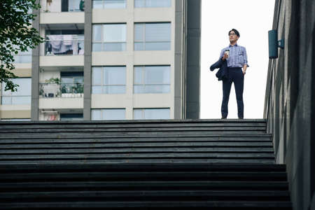 Confident ambitious Asian businessman standing on top of stairs with cup of coffee and jacket in hands and looking forwardの写真素材