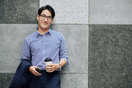 Portrait of handsome smiling young Asian man with cup of coffee and smartphone leaning on wall and looking at cameraの写真素材