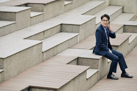 Smiling businessman resting on steps and talking on phone with coworkerの写真素材