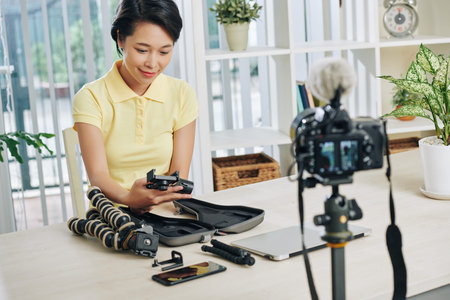 Smiling pretty young Asian woman shooting video review of tripods and monopods at home