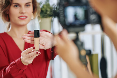 Positive beauty blogger showing bottle of new foundation when making review for her subscribers