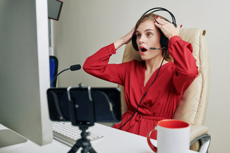 Shocked emotional streamer in headphones with microphone plying gmeale and shooting video at home
