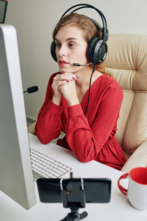 Female blogger in headphones with microphone watching video on computer screen and making comments