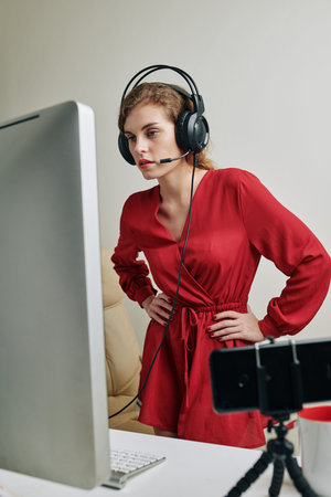Indignant pretty young woman in red dress and headphones with microphone watching stream and making...