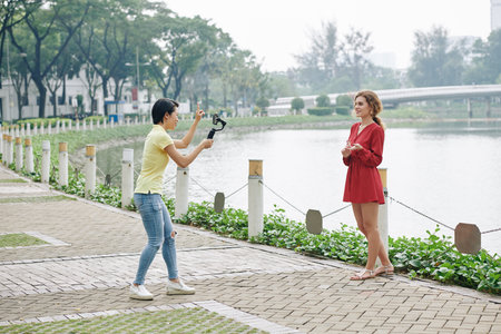 Two female friends shooting video on smartphone on bank of city river
