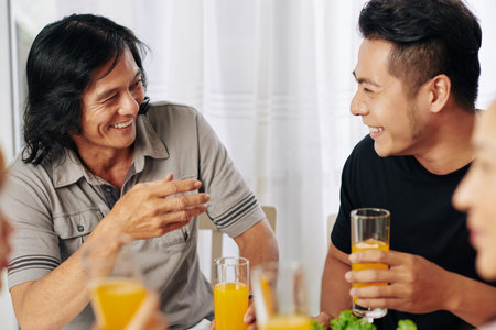Mature Asian man and his adult son drinking juice and joking around at family dinner