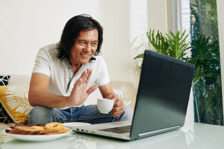 Positive mature Vietnamese man drinking coffee and waving with hand when video calling relative via...