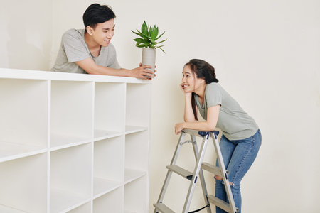 Pretty young Vietnamese woman standing on ladder and looking at her boyfriend standing at bookcase...