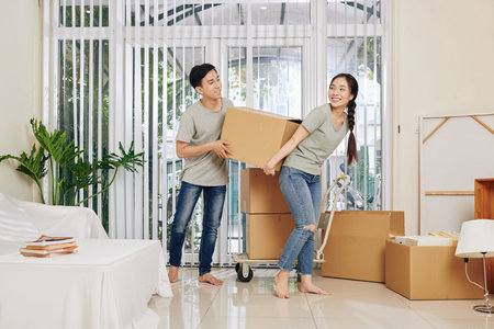 Happy young Vietnamese couple in casual clothes carrying heavy cardboard box when moving in new...