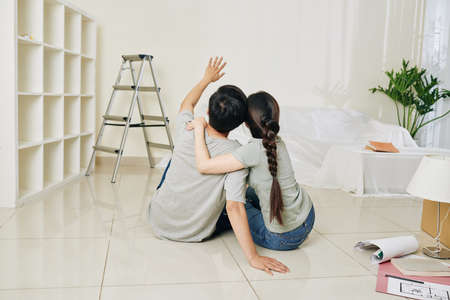Cheerful young hugging couple sitting on the floor in new apartment and discussing ideas for renovationの写真素材