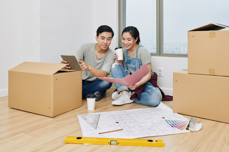 Cheerful young Vietnamese couple discussing construction plans in documents and on tablet computer...