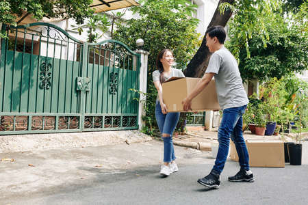 Happy young Vietnamese couple carrying heavy cardboard box to their new house when moving inの写真素材