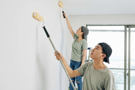 Young Asian professional house painters in caps using special rolls when painting room walls whiteの写真素材