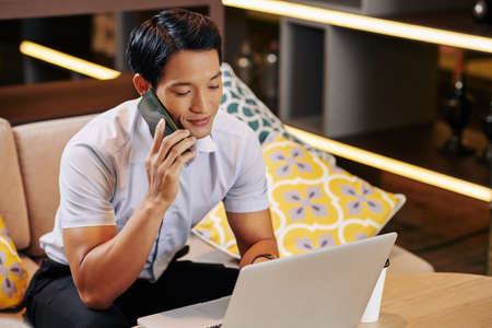 Young smiling Asian businessman calling on phone when reading document onlineの写真素材