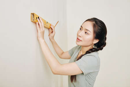 Lovely young Asian woman with spirit level drawing horizontal line on wall in her room when renovating houseの写真素材