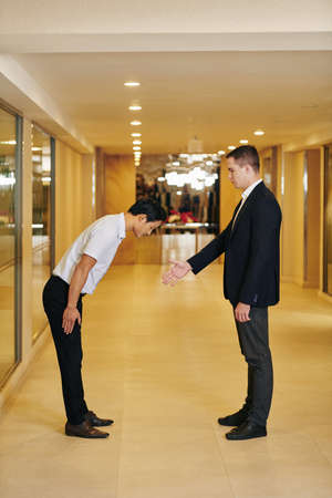 Caucasian businessman outstretching arm for handshake when his Asian colleague bending forward when greeting himの写真素材
