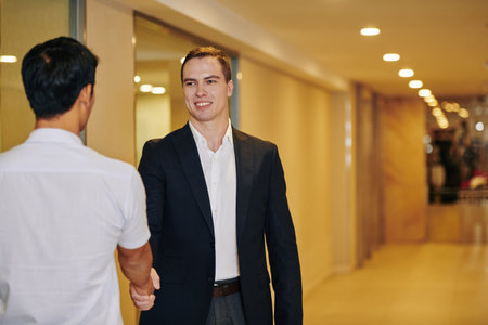 Handsome smiling successful businessman shaking hand of his new coworker when meeting him in office corridorの写真素材