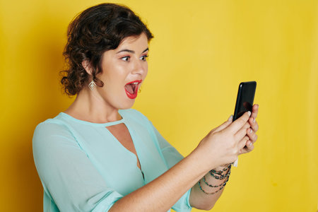 Amazed emotional young woman reading notification on her smartphone when standing against bright...