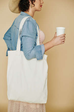 Cropped image of stylish young woman with linen reusable bag drinking paper glass of fresh waterの写真素材
