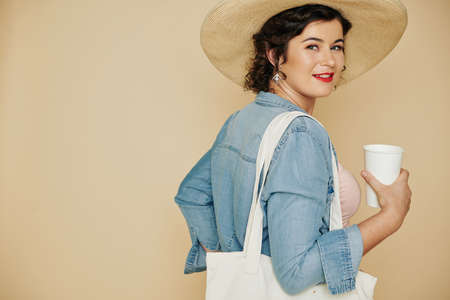 Portrait of young smiling woman in straw hat drinking cup of water and turning back to cameraの写真素材
