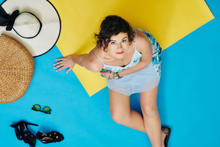 Beautiful woman sitting on yellow rug with hats and shoes around and looking at camera