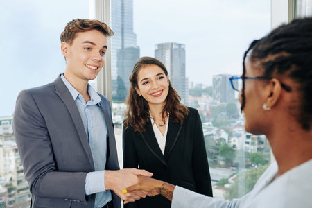 Cheerful young business people greeting new coworker in office and shaking her hand