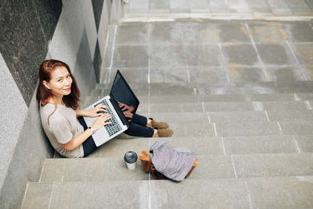 Positive female freelancer sitting on steps outdoors and coding on her laptopの写真素材